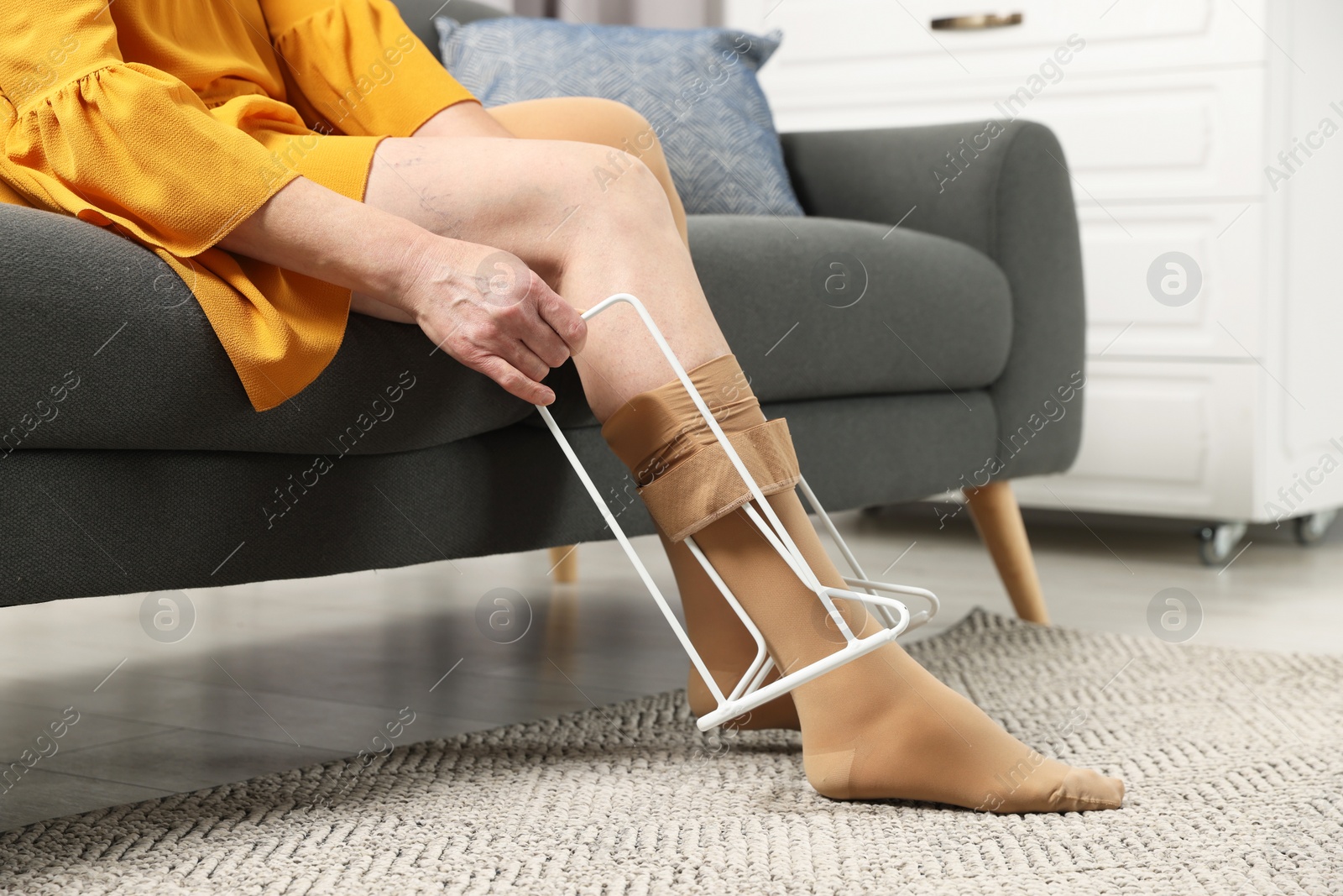 Photo of Woman putting on compression tights with stocking donner in living room, closeup. Prevention of varicose veins