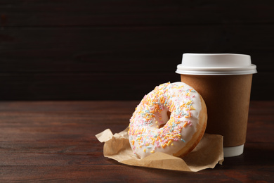 Photo of Yummy donut and paper cup on wooden table against brown background, space for text