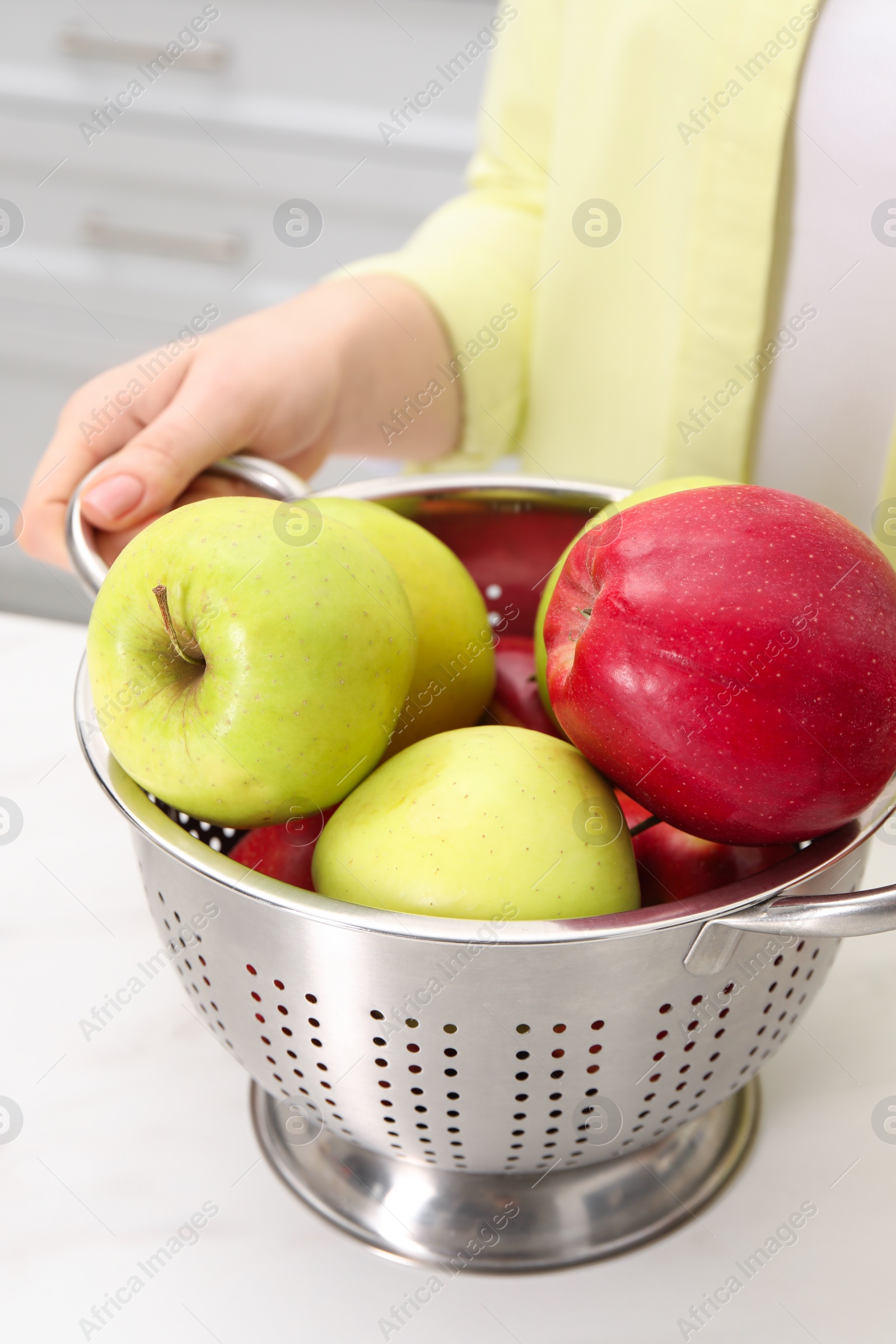 Photo of Woman holding colander with fresh apples at white marble table in kitchen, closeup