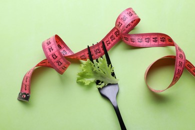 Photo of Fork with measuring tape and lettuce leaf on light green background, flat lay. Diet concept