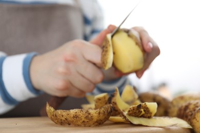 Photo of Woman peeling fresh potato with knife at table, focus on peels