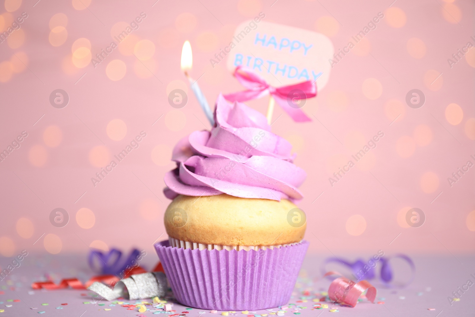 Photo of Beautiful birthday cupcake, streamers and confetti against pink background with blurred lights