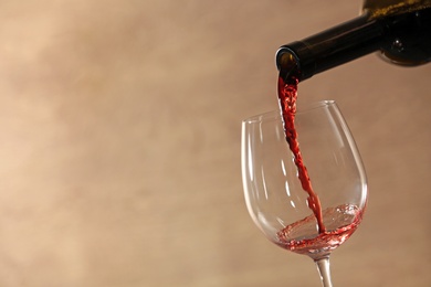 Photo of Pouring red wine into glass from bottle against blurred beige background, closeup. Space for text