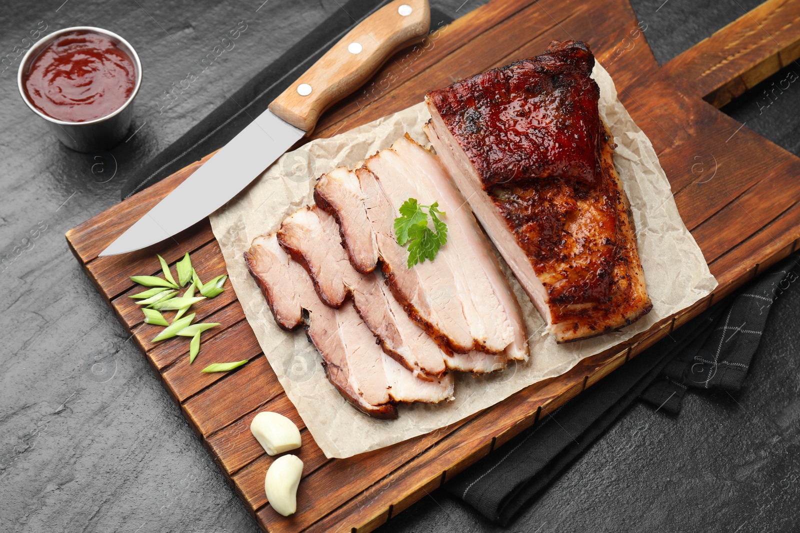 Photo of Pieces of baked pork belly served with sauce and parsley on black textured table, above view