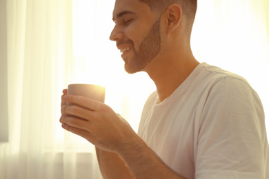 Photo of Young man with cup of coffee near window at home, closeup. Lazy morning
