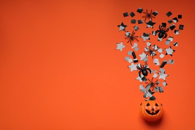 Photo of Plastic pumpkin basket, black confetti, spiders and space for text on orange background, flat lay. Halloween celebration