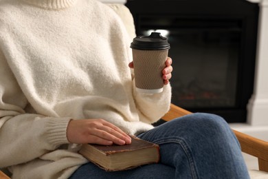 Photo of Woman holding takeaway cardboard cup and book indoors, closeup. Coffee to go