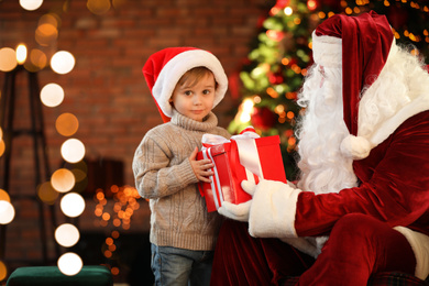 Photo of Santa Claus and little boy with gift near Christmas tree indoors