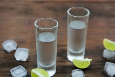 Photo of Mexican tequila shots with lime slices and ice cubes on wooden table, closeup. Drink made from agave