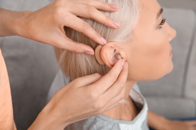 Photo of Young woman putting hearing aid in mother's ear indoors, closeup