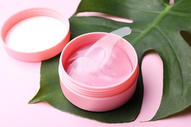 Photo of Under eye patches in jar with spatula and green leaf on light pink background. Cosmetic product