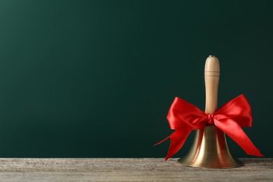 Photo of Golden bell with red bow on wooden table near green chalkboard, space for text. School days