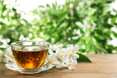 Photo of Glass cup of aromatic jasmine tea and fresh flowers on wooden table, space for text