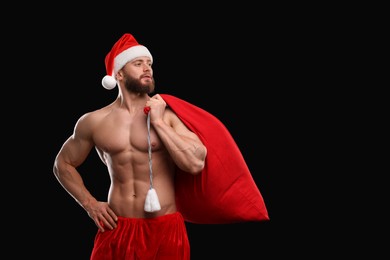 Photo of Muscular young man in Santa hat holding bag with presents on black background, space for text