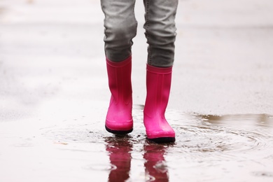 Photo of Little girl wearing rubber boots splashing in puddle on rainy day, focus of legs. Autumn walk
