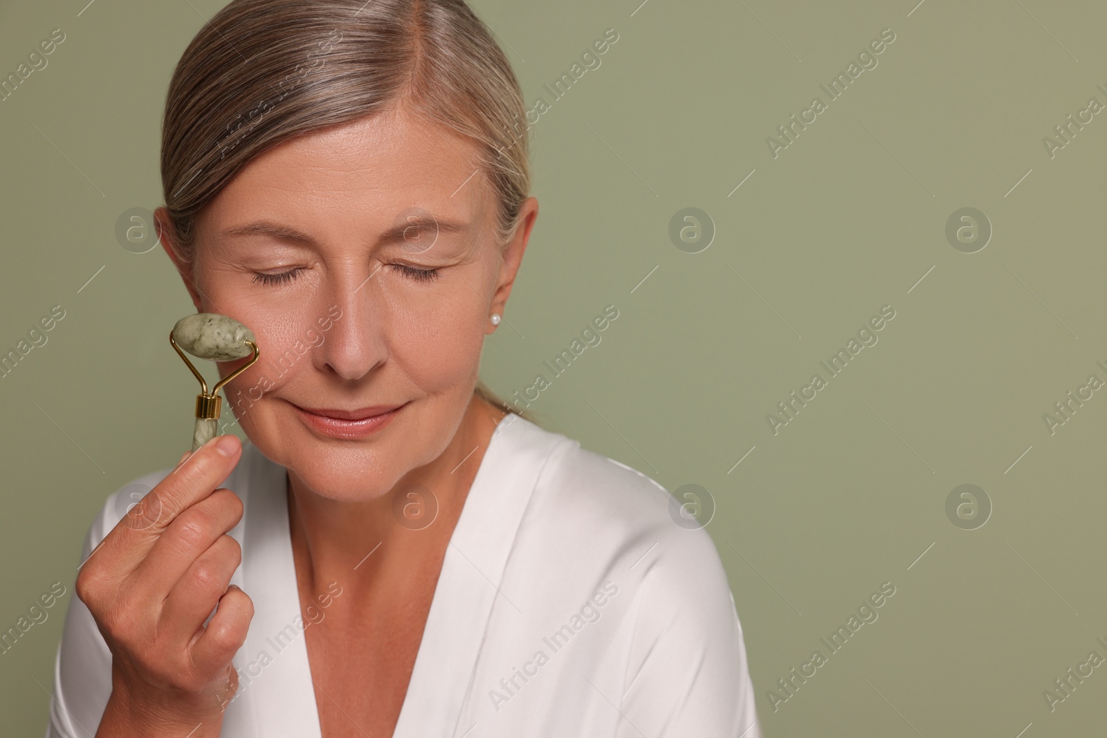 Photo of Woman massaging her face with jade roller on green background, space for text