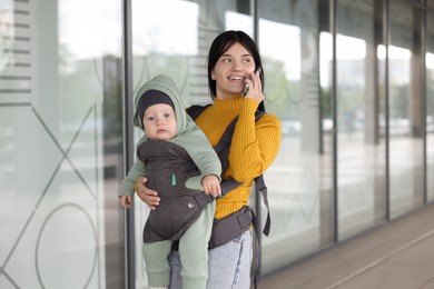 Photo of Mother holding her child in sling (baby carrier) while talking on smartphone near building outdoors