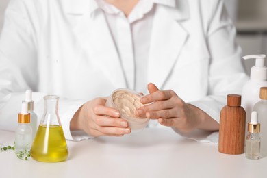 Photo of Dermatologist with jar testing cosmetic product at white table indoors, selective focus