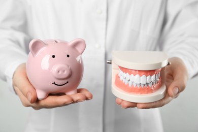 Photo of Dentist holding educational typodont model and piggy bank on light background, closeup. Expensive treatment
