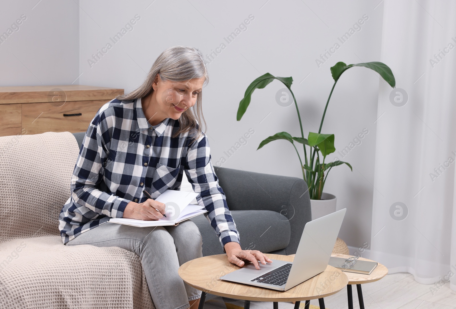 Photo of Beautiful senior woman writing something in notebook while using laptop at home