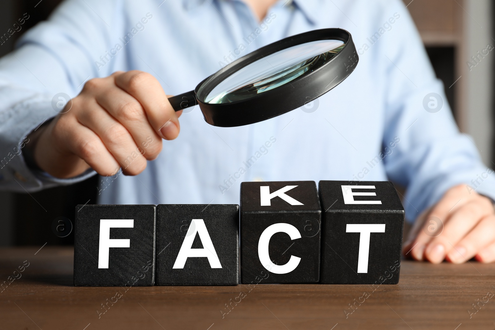 Photo of Woman holding magnifying glass above words FAKE and FACT made with black cubes at wooden table, closeup