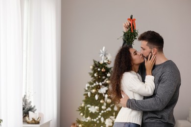 Photo of Happy couple kissing under mistletoe bunch in room decorated for Christmas