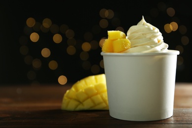 Cup of tasty frozen yogurt with mango on wooden table against blurred lights. Space for text