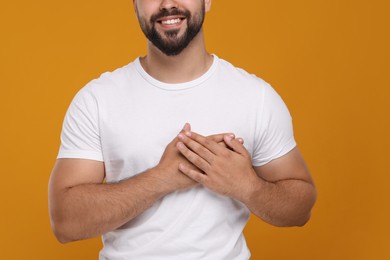 Photo of Thank you gesture. Grateful man holding hands near heart on orange background, closeup