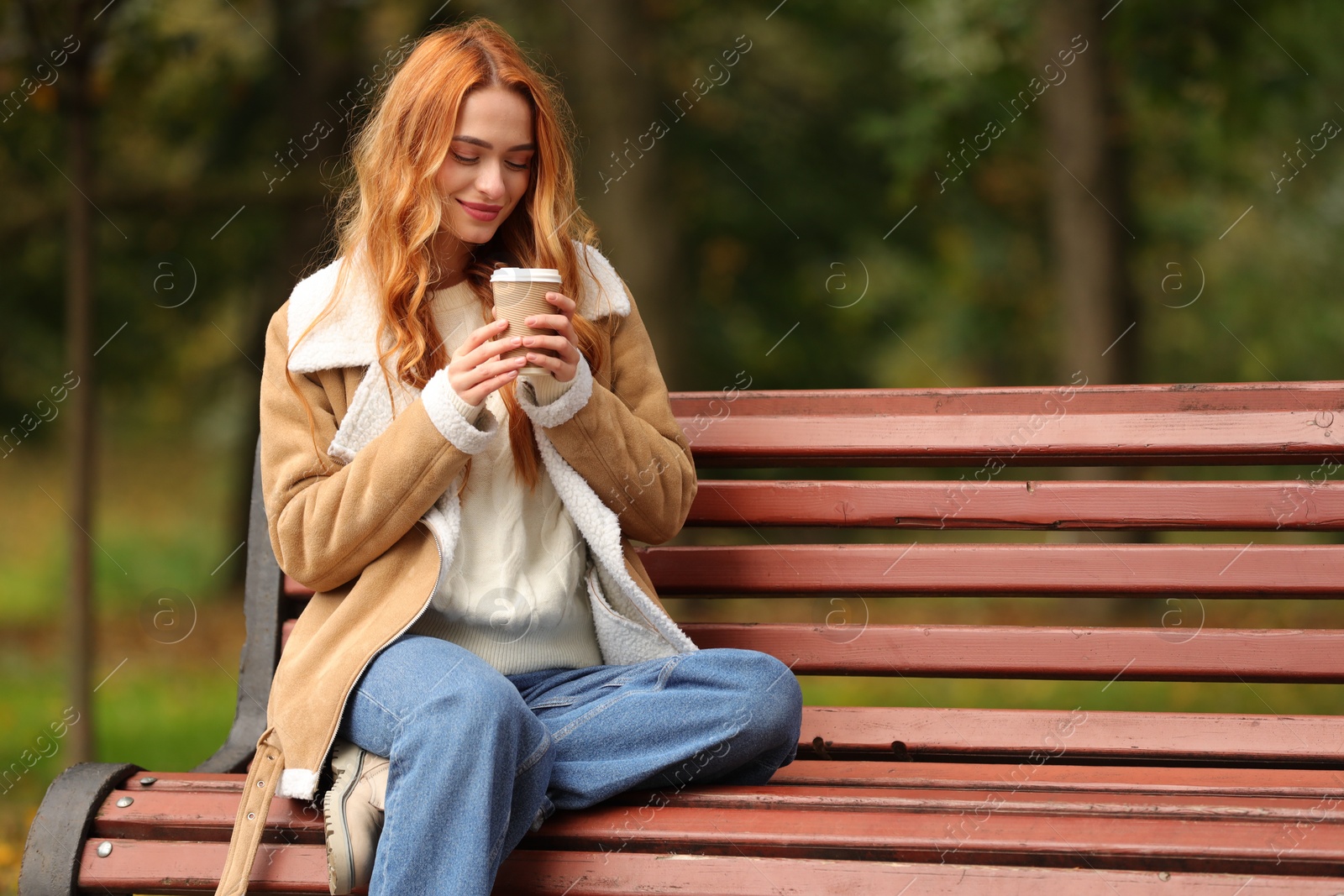Photo of Beautiful woman with paper cup sitting on bench in autumn park. Space for text