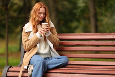 Beautiful woman with paper cup sitting on bench in autumn park. Space for text