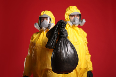 Photo of Man and woman in chemical protective suits holding trash bag on red background. Virus research