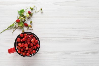 Fresh wild strawberries in mug near stems with flowers and leaves on white wooden table, flat lay. Space for text