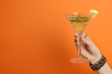 Photo of Woman holding martini glass of refreshing cocktail with lemon slice and rosemary on orange background, closeup. Space for text