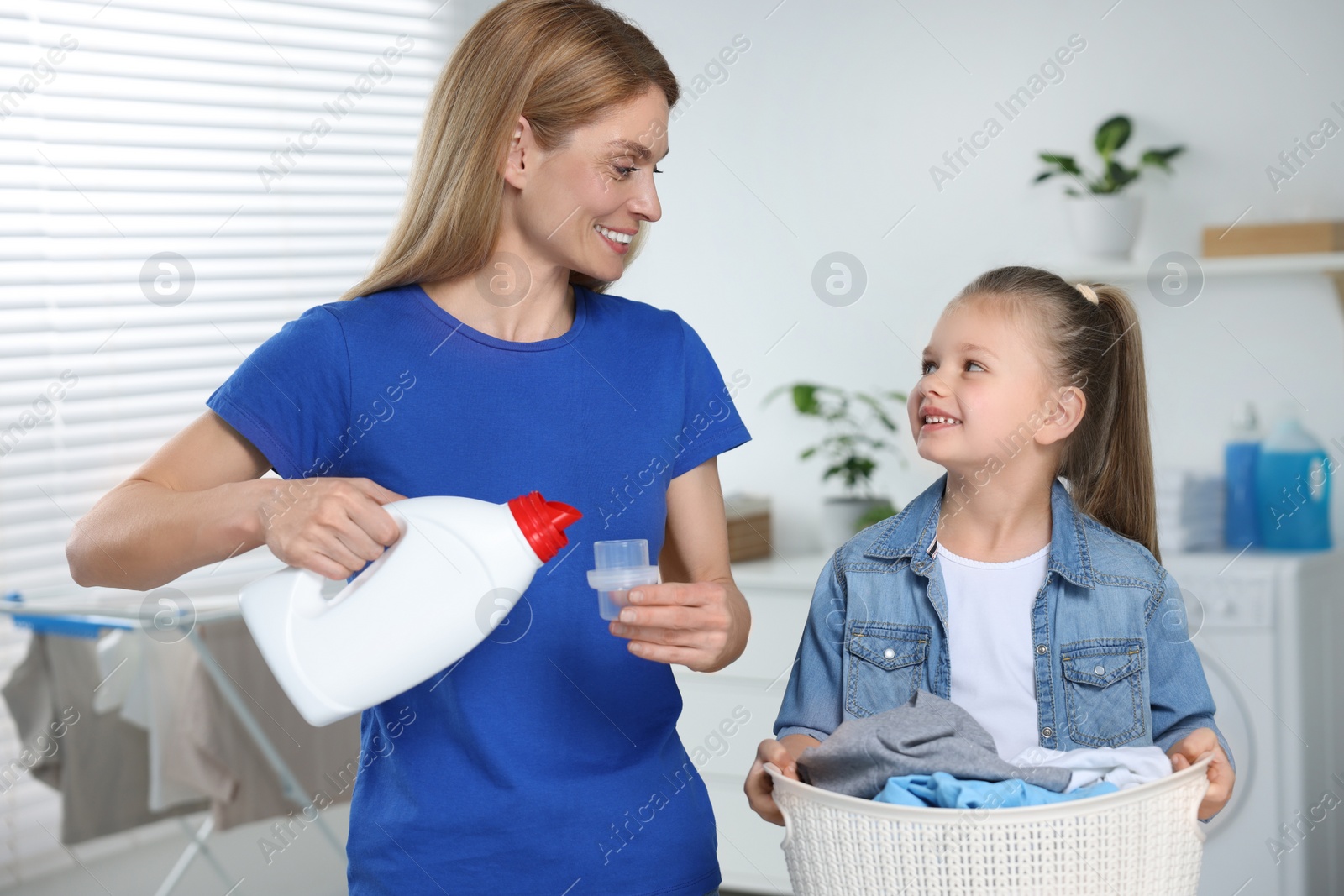 Photo of Mother pouring fabric softener from bottle into cap and daughter holding basket with dirty clothes in bathroom