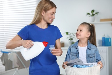 Photo of Mother pouring fabric softener from bottle into cap and daughter holding basket with dirty clothes in bathroom