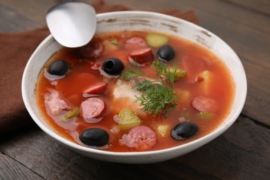 Photo of Meat solyanka soup with thin dry smoked sausages in bowl on wooden table, closeup