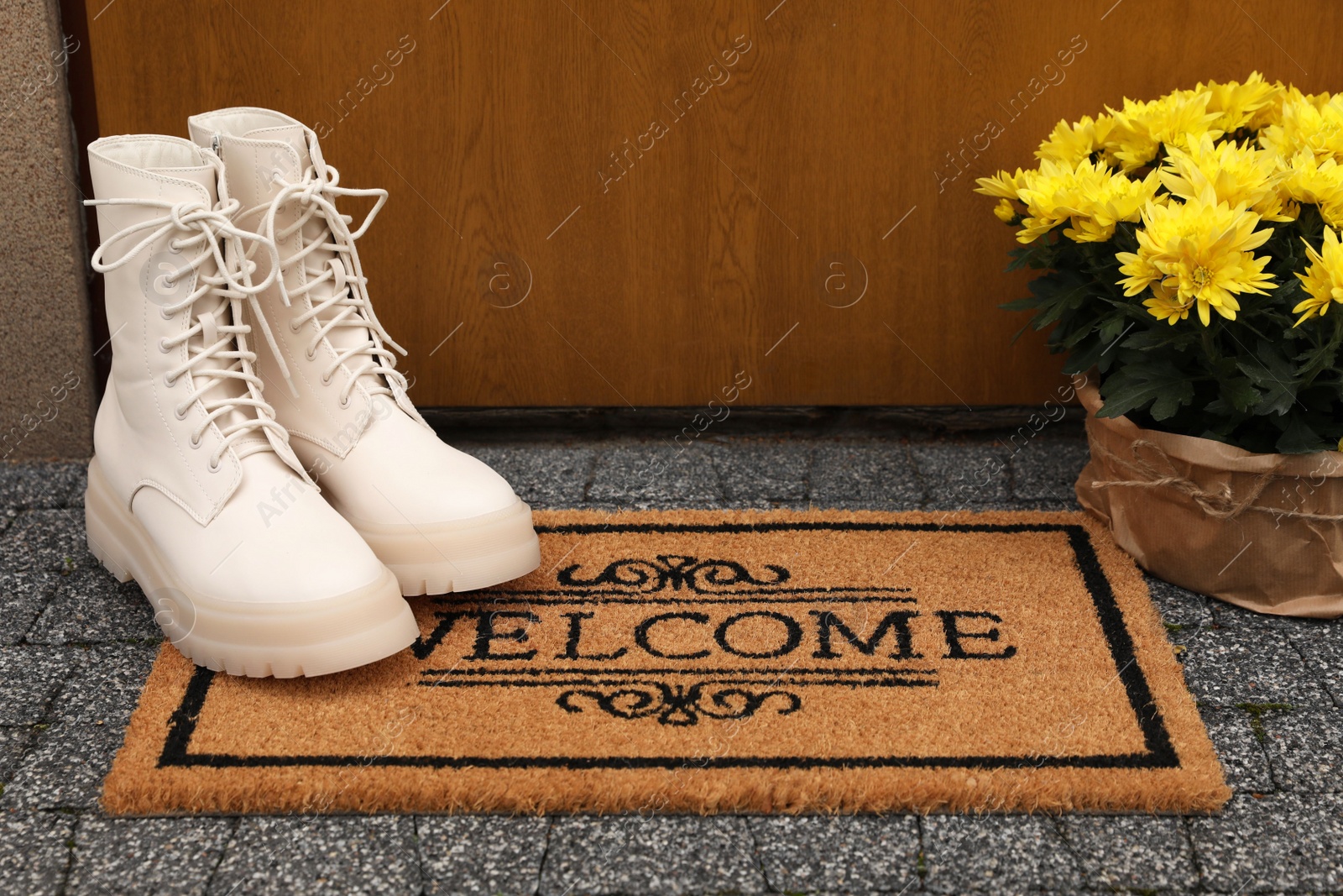 Photo of Doormat with word Welcome, stylish boots and beautiful flowers on floor near entrance