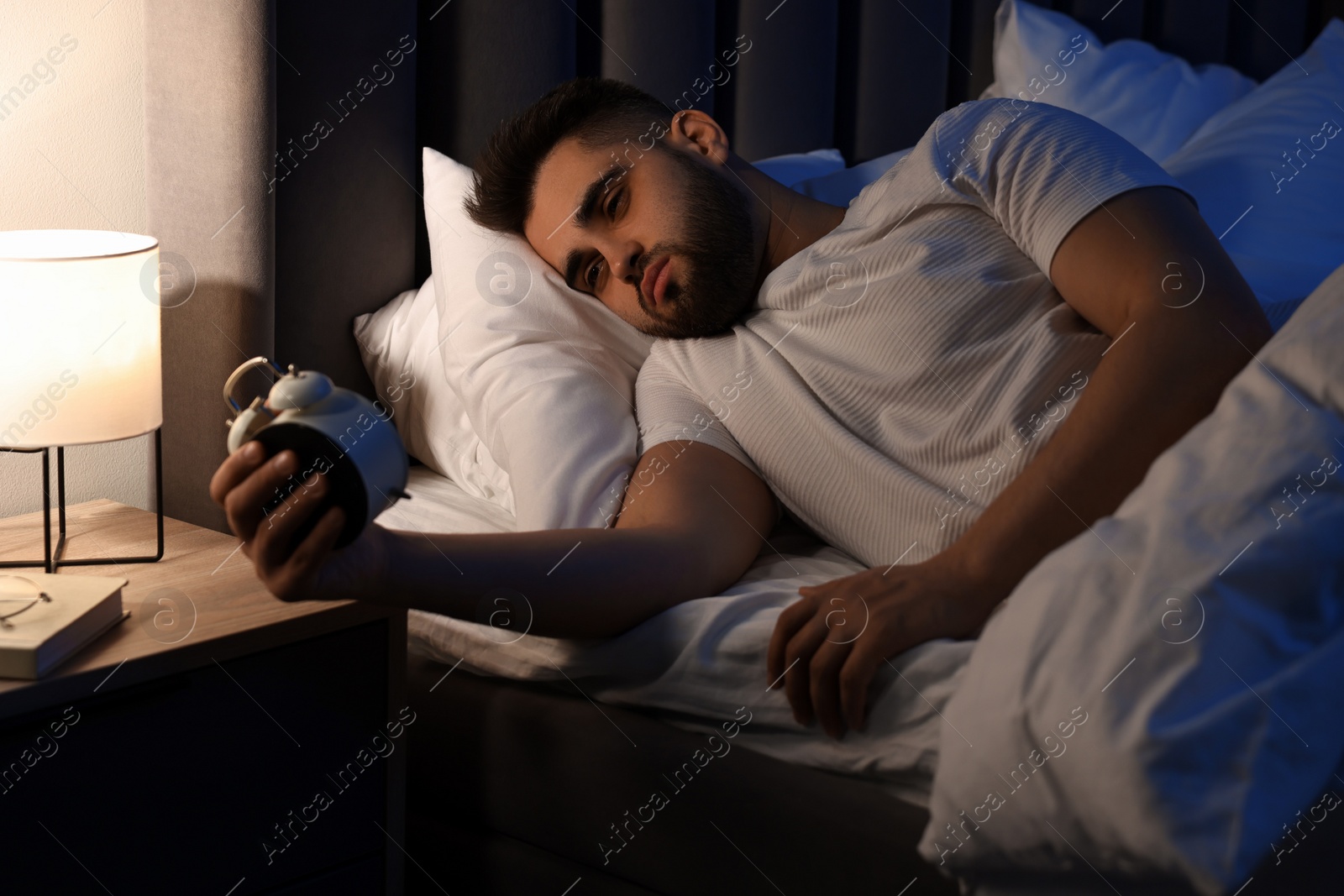 Photo of Frustrated man with alarm clock on bed. Insomnia problem