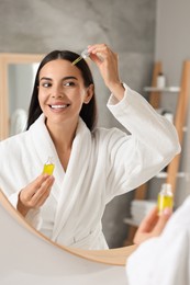 Beautiful young woman applying serum onto her face in bathroom