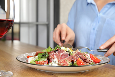 Photo of Woman eating delicious prosciutto salad with vegetables at wooden table indoors, closeup