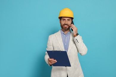 Photo of Professional engineer in hard hat with clipboard talking on phone against light blue background