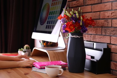 Photo of Stylish vase with fresh flowers and woman working on computer at wooden table, closeup