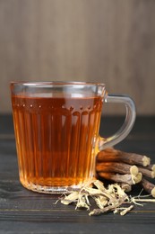 Photo of Aromatic licorice tea in cup and dried sticks of licorice root on black wooden table, closeup