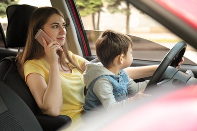 Photo of Mother with little son on knees driving car and talking by phone. Child in danger