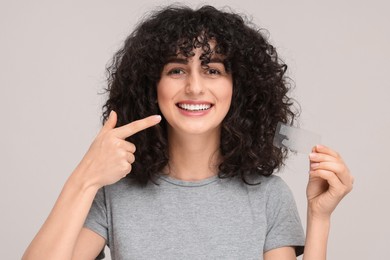 Photo of Young woman holding whitening strips and pointing at her teeth on light grey background