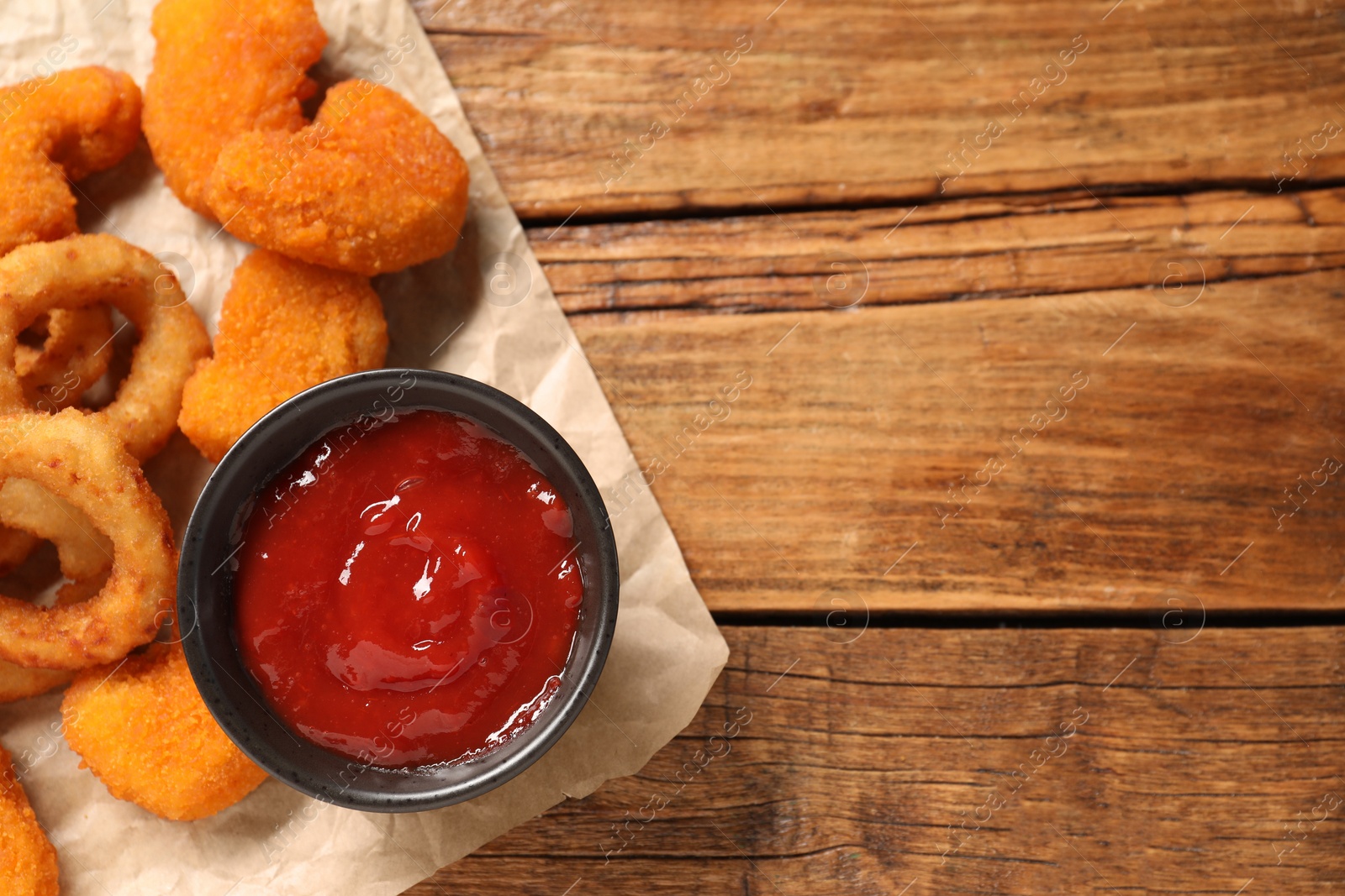 Photo of Tasty fried onion rings, chicken nuggets and ketchup on wooden table, top view. Space for text