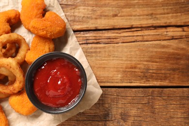 Tasty fried onion rings, chicken nuggets and ketchup on wooden table, top view. Space for text