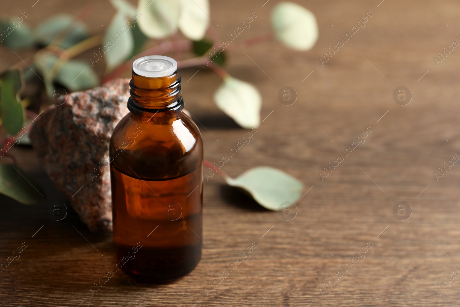 Photo of Bottle of eucalyptus essential oil, stone and leaves on wooden table. Space for text