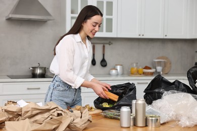 Photo of Garbage sorting. Woman putting food waste into plastic bag at table in kitchen