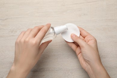Photo of Woman pouring micellar water from bottle onto cotton pad at wooden table, top view